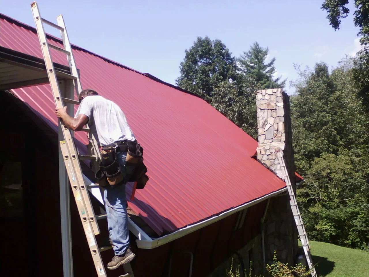 Roofer on a ladder beside a red metal roof and stone chimney — Soffit & Fascia Repair by Roofing Brothers in Mountlake Terrace